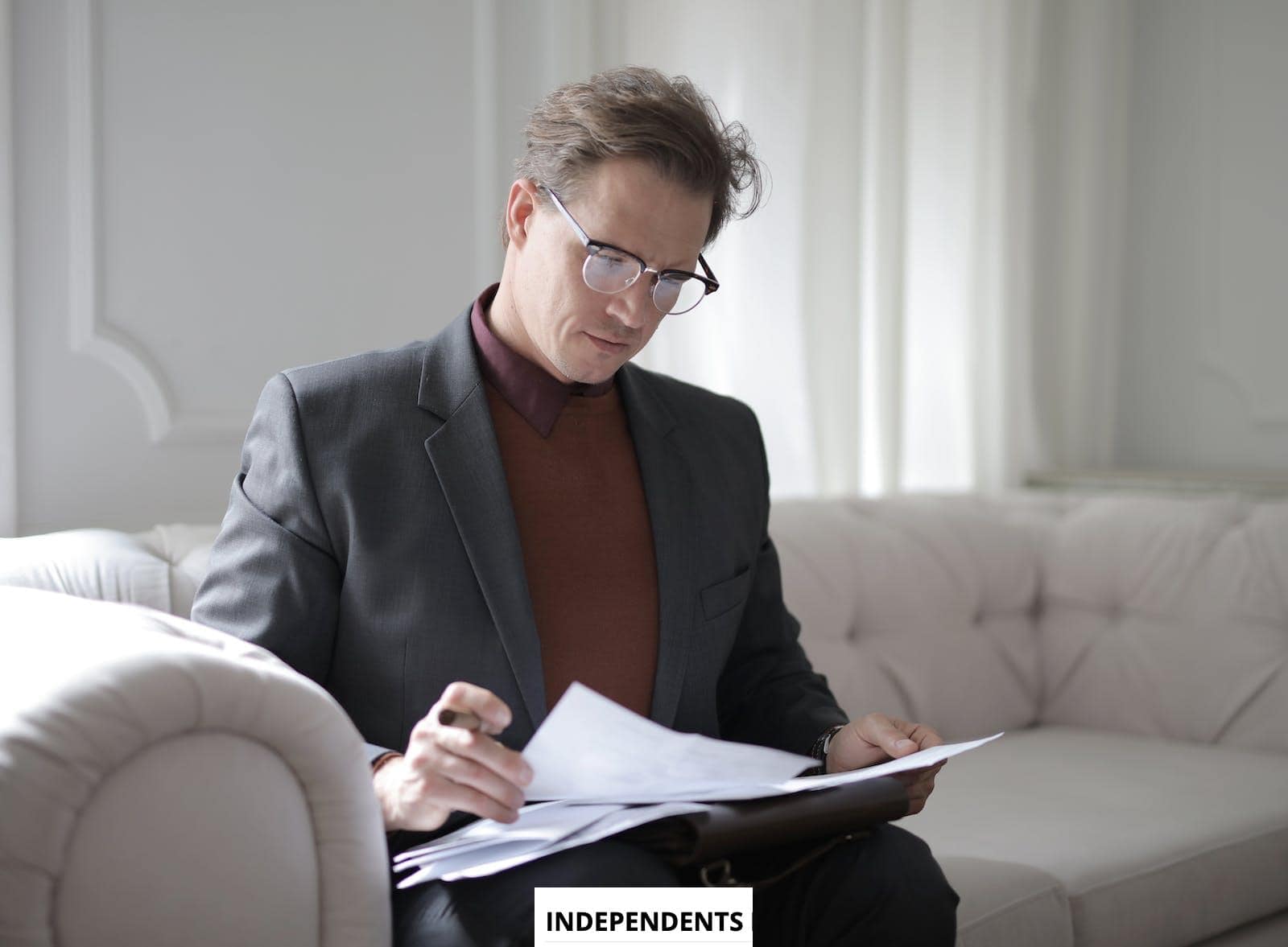 Elegant adult man in jacket and glasses looking through documents while sitting on white sofa in luxury room
